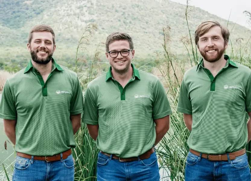 Three Agrigistics founders in green branded shirts standing outdoors in an agricultural field setting